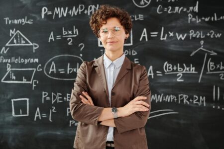 Uśmiechnięta nauczycielka w okularach stoi z założonymi rękami na tle czarnej tablicy zapisanej białymi wzorami geometrycznymi i algebraicznymi. Widoczne są rysunki trójkąta, równoległoboku oraz wzory na pole i obwód figur, które ułatwiają przygotowanie do matury z matematyki. Więcej informacji o skutecznych kursach maturalnych z matematyki znajdziesz na stronie Centrum Edukacyjnego Symposio.